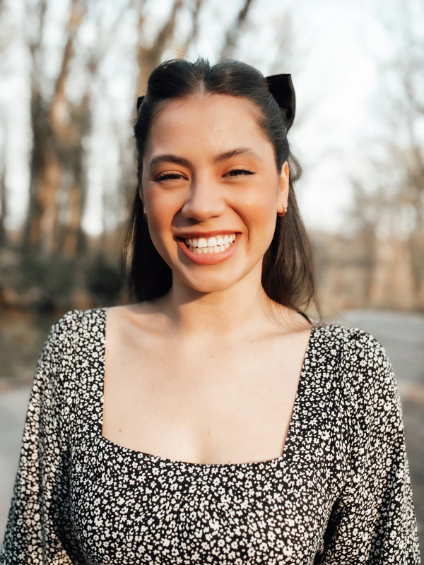 A young woman with dark hair, smiling broadly with her eyes closed, standing outdoors with blurred trees in the background.