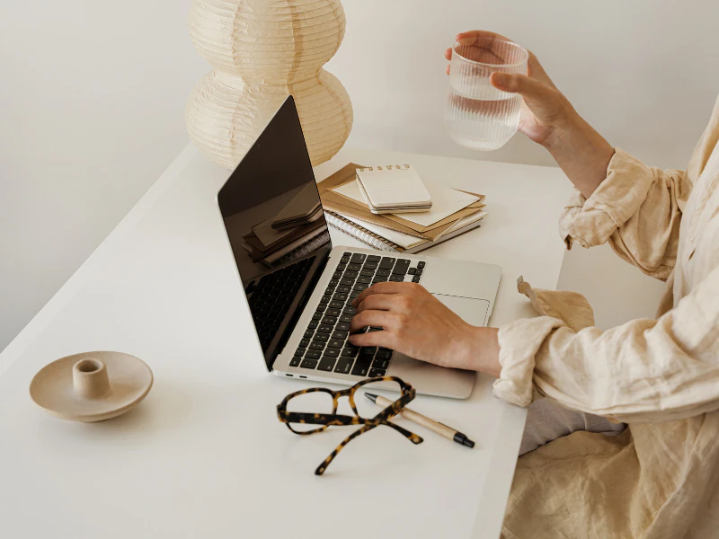 A person working at a white desk with a laptop, notebooks, a pair of glasses, a glass of water, a ceramic vase, and a paper lantern.