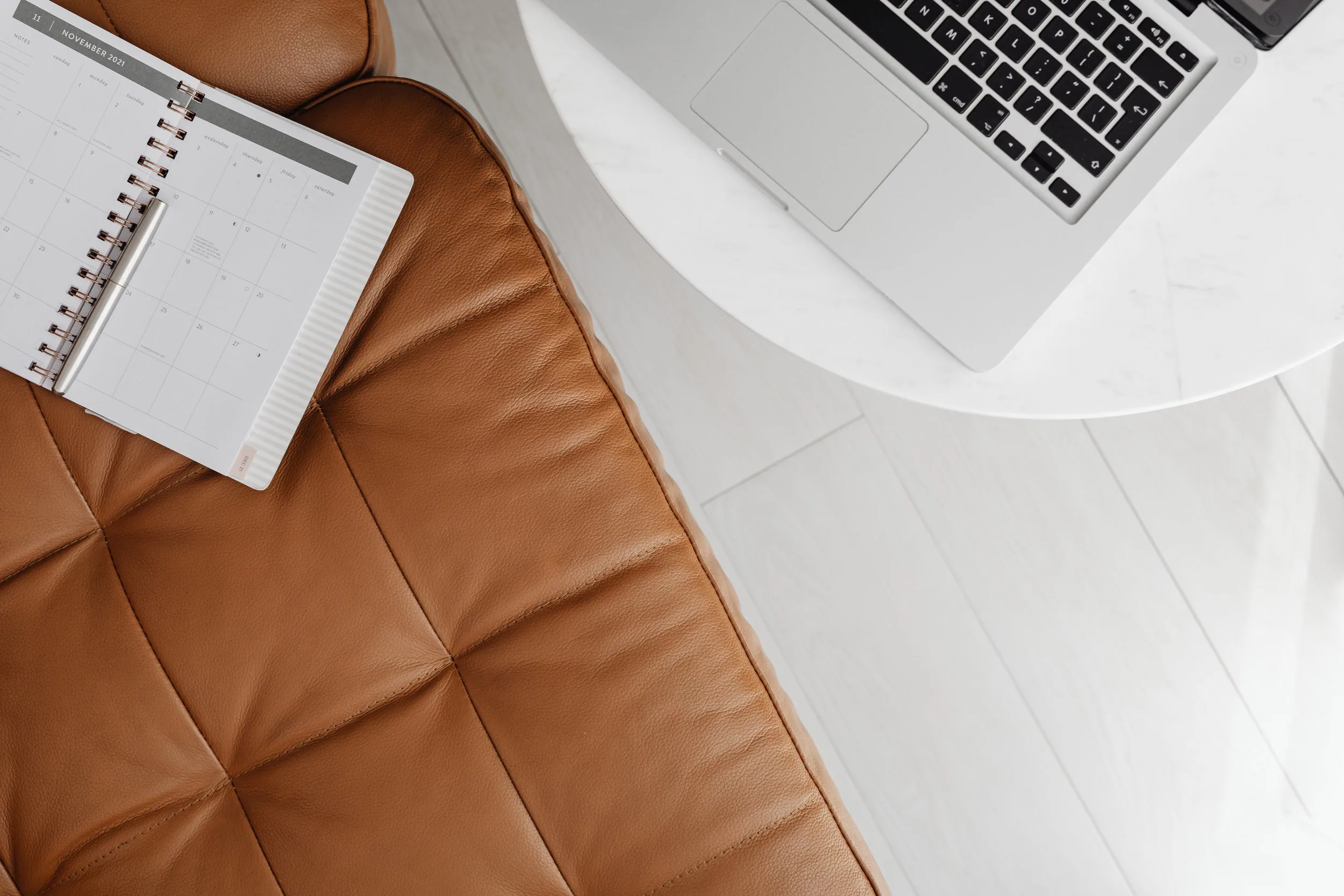 A brown leather tufted sofa with a white marble-top table nearby. On the sofa, there is a planner with a silver pen on top. A laptop with a black keyboard is open on the table.