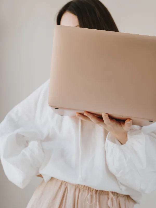 Person holding a closed beige laptop in front of their face, wearing a white jacket and beige pants.