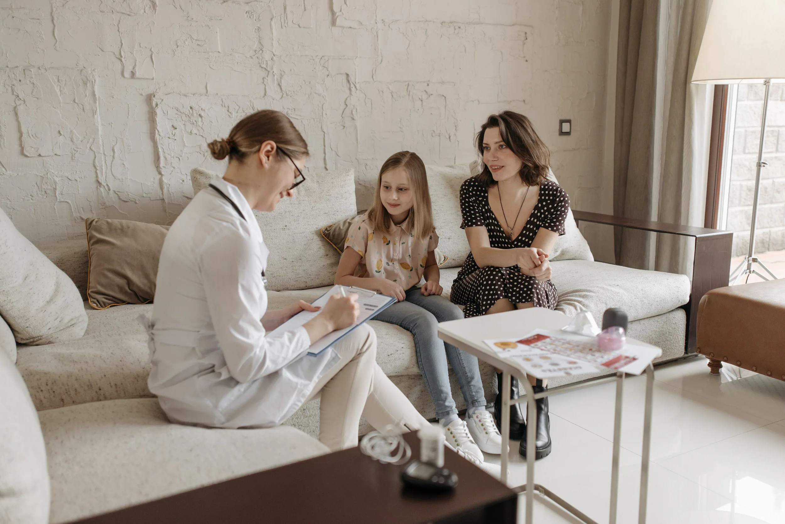 A woman in a white coat, presumably a doctor, consulting with a young girl and another woman in a cozy living room with a white brick wall. The doctor is taking notes, the girl is seated between the women, and they are all engaged in conversation. There are cushions on the sofa, a white table with some papers, and a floor lamp near a large window with curtains.