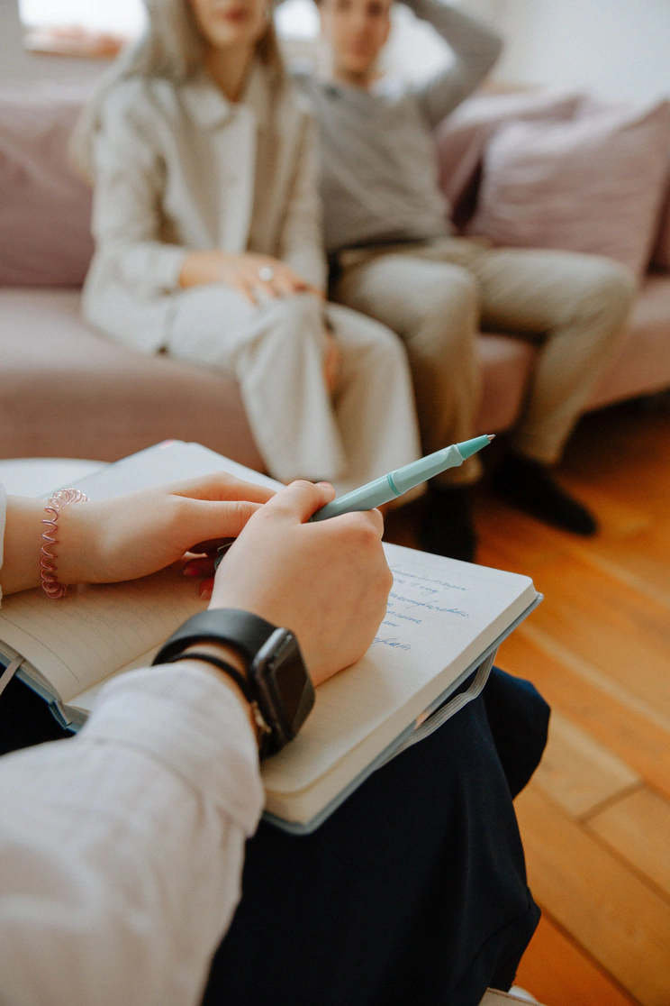 A person with a bracelet on their wrist taking notes in a notebook. In the background, two people sitting on a couch, one man and one woman, are blurred out.