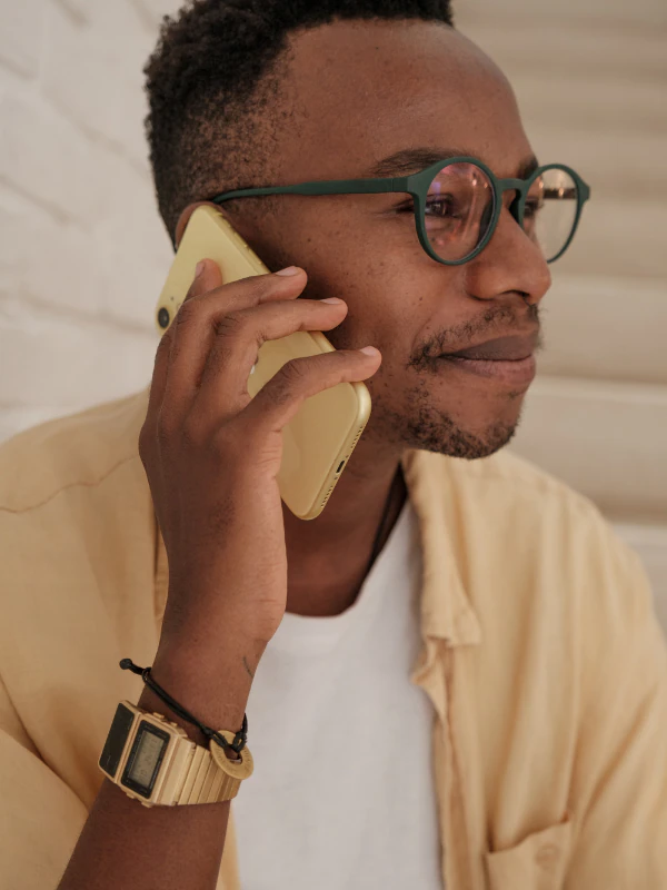 Man with glasses talking on a yellow smartphone, wearing a beige shirt, white t-shirt, bracelet, and watch.