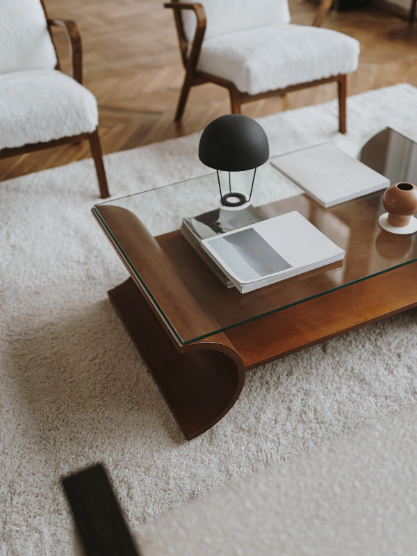 A glass coffee table with a wooden base, placed on a white shag rug in a living room. The table has a black desk lamp, a stack of magazines, some white books, and a small brown vase.